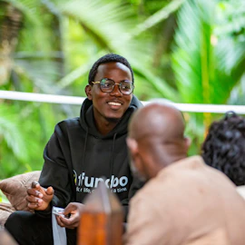 A man is in conversation with a group of other people. He is a Black man with short black hair, wearing glasses and a black hoodie. He is smiling.